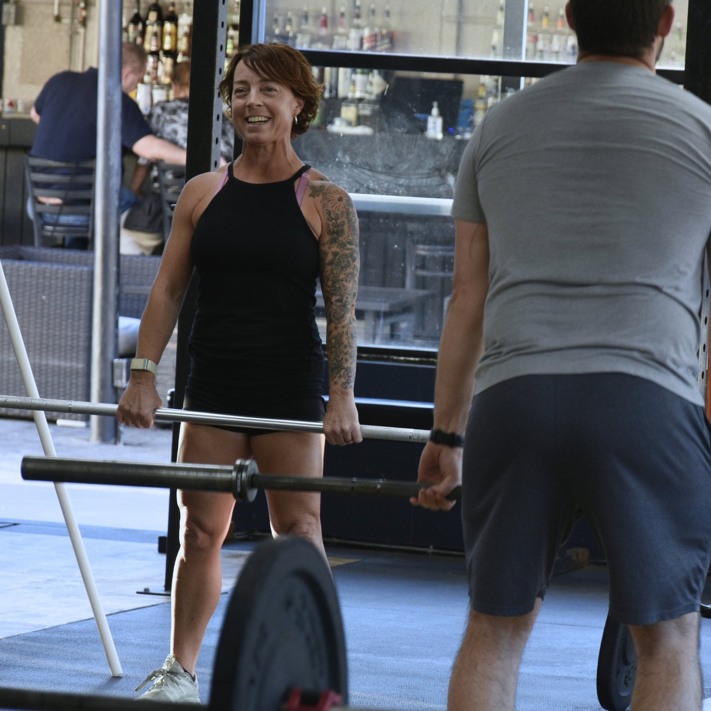 Stacy coaching an athlete through a barbell lift, smiling across the bar